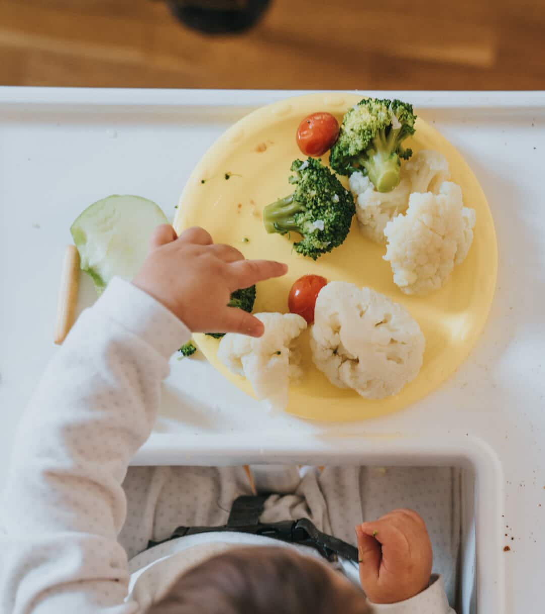 Baby in a high chair eating broccoli, cauliflower and tomatoes 